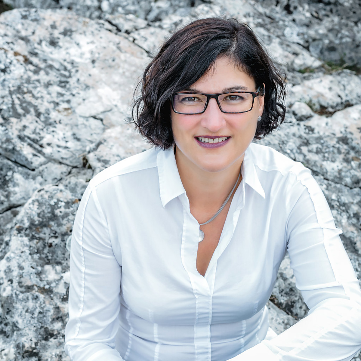 Close-up of Tanja Reischl-Stenske, outdoors sitting on a rock, in a white shirt, smiling.